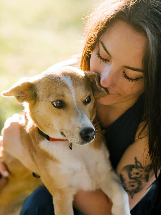 Brunette girl with tattoo on shoulder hugs dog.