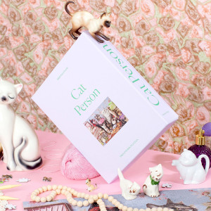 Portrait of a light purple puzzle box that reads "Cat Person" with a pink patterned wallpaper and various cat figurines