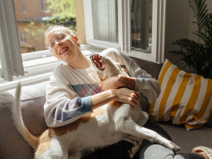 Girl sitting on couch hugging her playful dog with the window open to outside