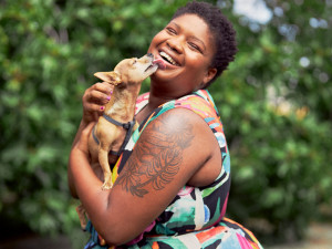 African American woman with floral arm tattoos and short curly hair laughing with joy while holding her small Chihuahua dog outside